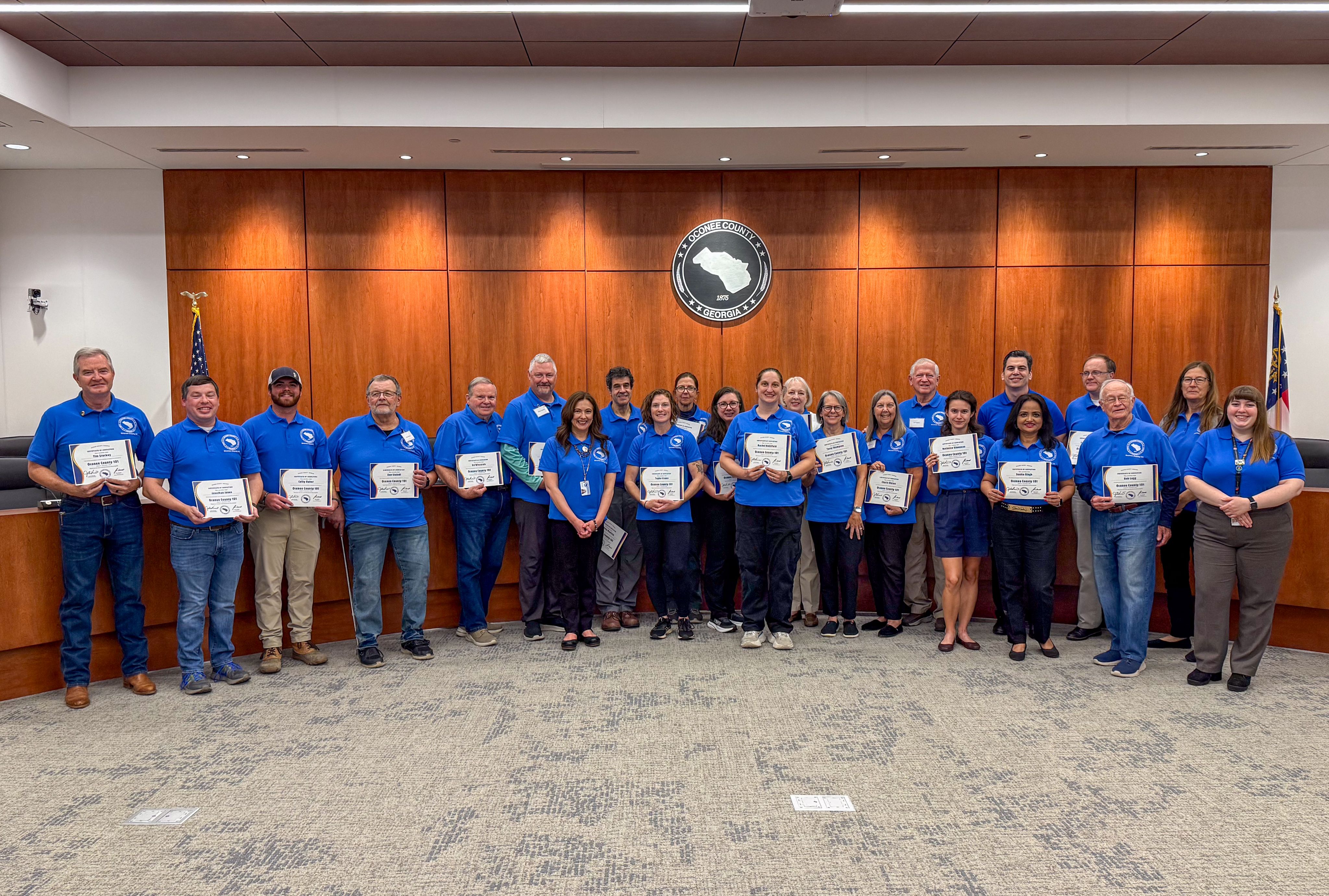 Oconee County 101 graduates pose in the BOC Chamber with their certificates wearing OC 101 blue polo shirts